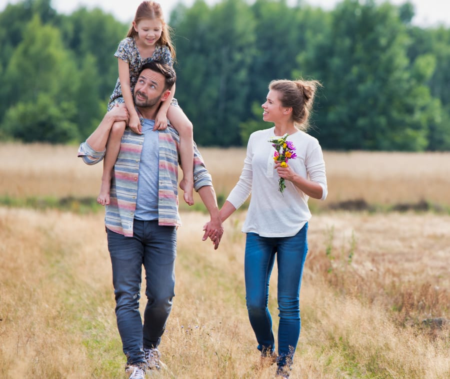 Family of four smiling and laughing together outdoors as the father carries a young boy on his shoulders.
