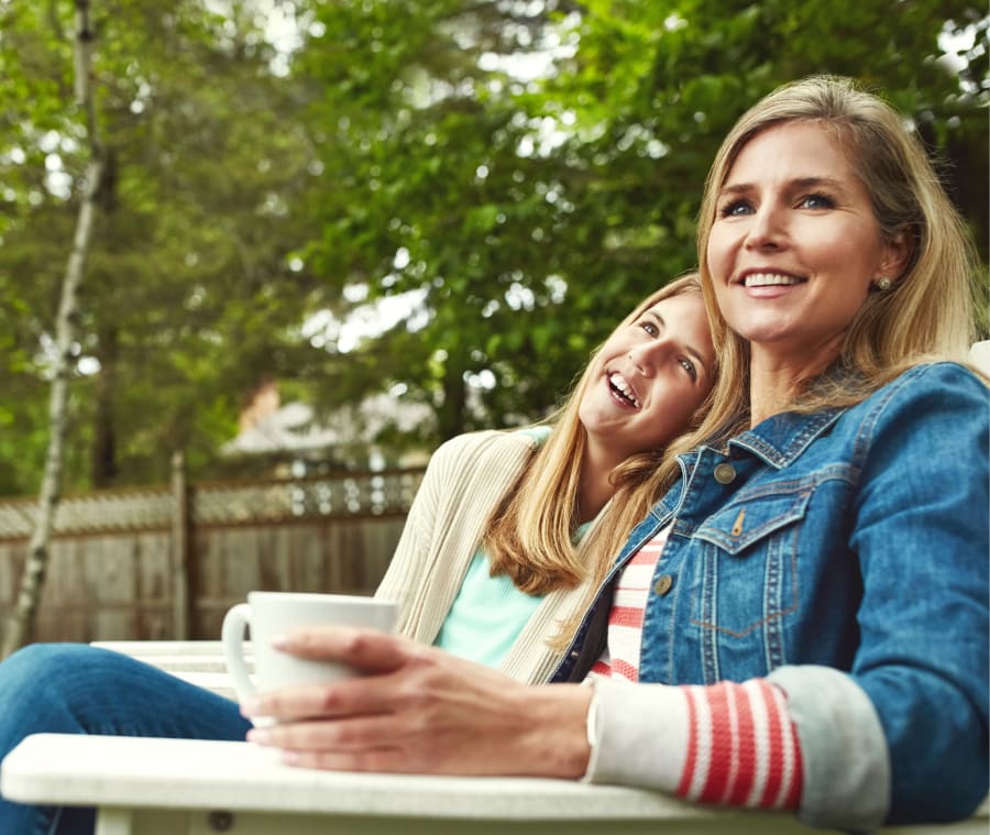 Mother and daughter sitting outdoors together, with the daughter resting her head on her mother's shoulder while her mother enjoys a cup of coffee.