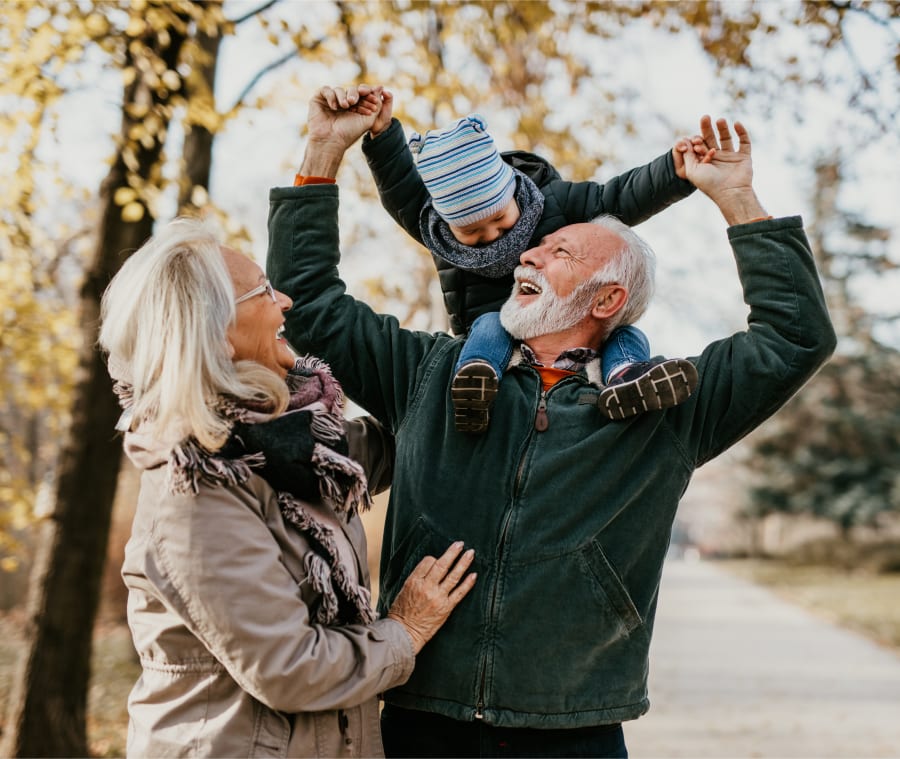 Elderly woman and man smiling with their grandson, as the young boy sits on his grandfather's shoulders.