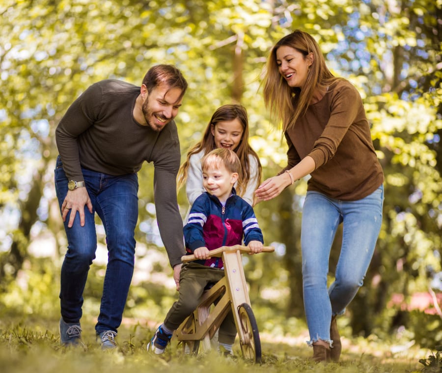 Family of four enjoying time outdoors in the woods, with a young boy riding his bike while the family walks and laughs together.