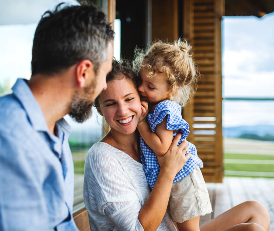 Family of three smiling together as a young girl kisses her mother on the cheek.